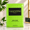 A 100 ml box of Valentino Donna Born in Roma Green Stravaganza Eau de Parfum, featuring a bright lime green geometric pattern with a black central label. The box is displayed on a marble surface with a blurred background containing a plant and a gold-framed object.