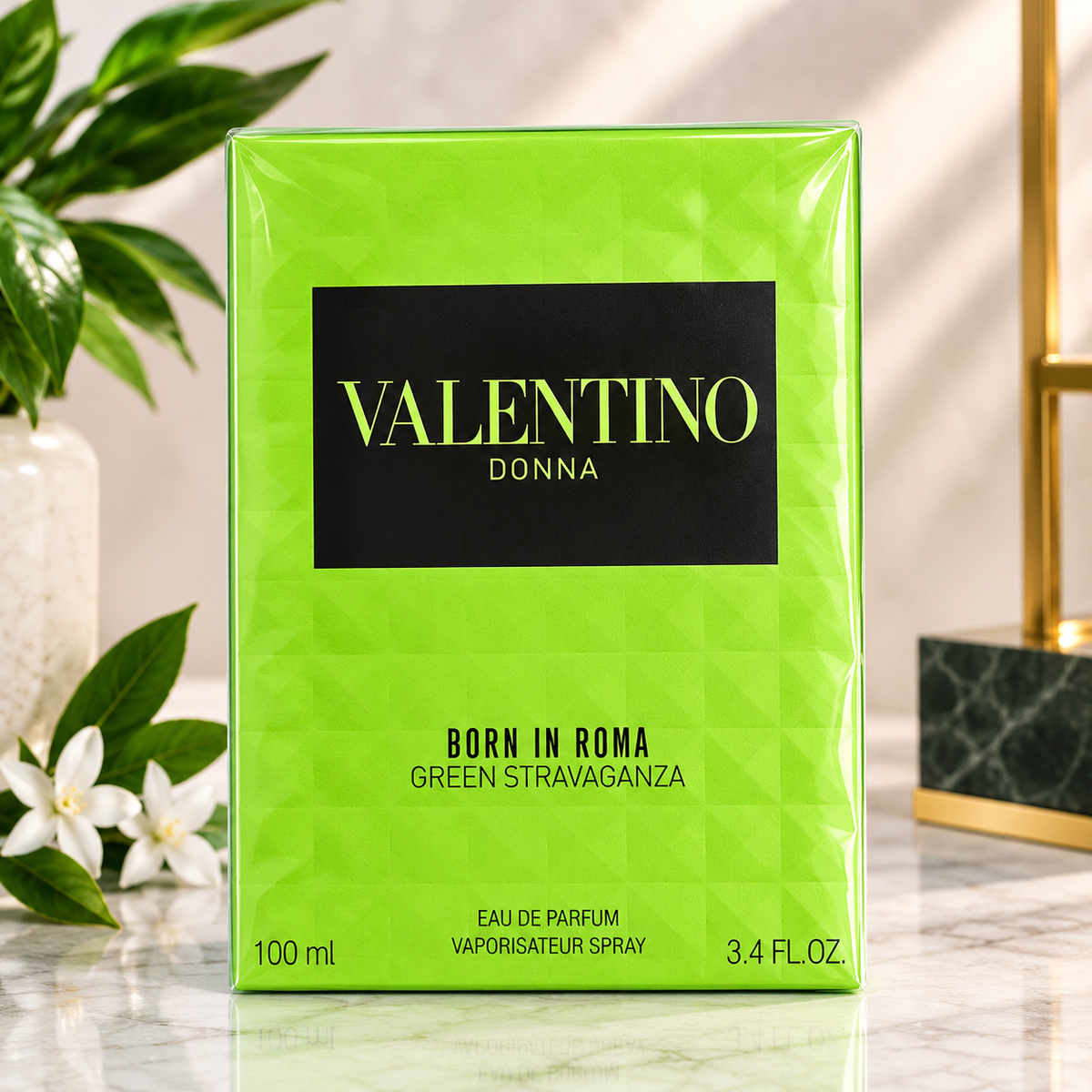 A 100 ml box of Valentino Donna Born in Roma Green Stravaganza Eau de Parfum, featuring a bright lime green geometric pattern with a black central label. The box is displayed on a marble surface with a blurred background containing a plant and a gold-framed object.