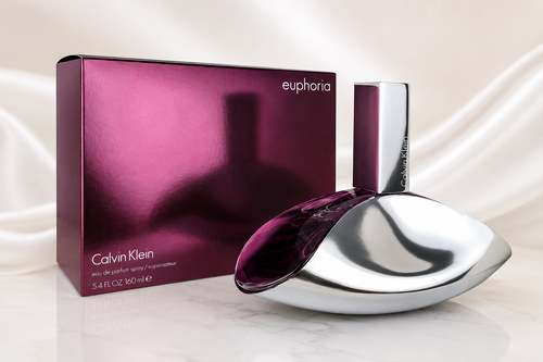 Calvin Klein Euphoria Eau de Parfum bottle with a reflective silver cap and dark purple glass, shown next to its magenta box on a light marble surface with draped satin fabric in the background.