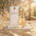 Dior J'adore Eau de Parfum bottle and its white box, styled on a stone slab with white flowers and olive branches against a warm beige stone wall background.