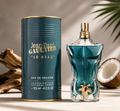 Jean Paul Gaultier Le Beau Eau de Toilette bottle and its cylindrical packaging box, styled on a warm beige surface with decorative driftwood and a coconut shell in the background.